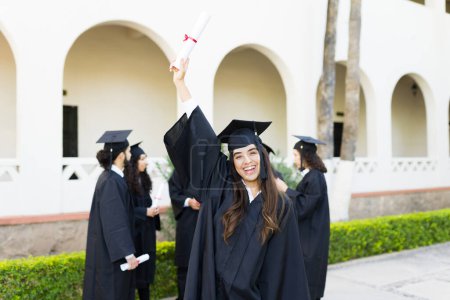 Téléchargez les photos : Jeune femme excitée montrant son diplôme universitaire et célébrant après avoir assisté à sa cérémonie de remise des diplômes au collège - en image libre de droit