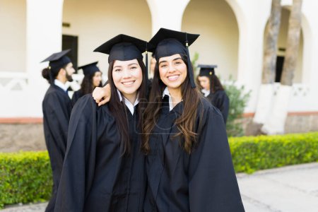 Téléchargez les photos : Femmes heureuses meilleurs amis étreignant et souriant portant une robe de graduation et casquette alors qu'à leur université - en image libre de droit