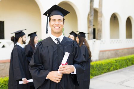 Téléchargez les photos : Attrayant homme hispanique souriant regardant heureux tenant son diplôme universitaire lors de sa cérémonie de remise des diplômes - en image libre de droit