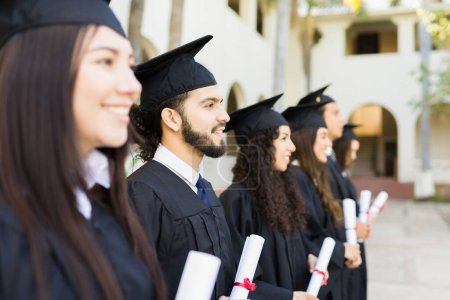 Foto de Grupo de graduados universitarios posando recibir su diploma universitario y sonriendo con vestidos de graduación y gorras - Imagen libre de derechos
