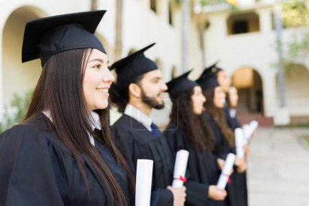 Téléchargez les photos : Diplômés d'université souriant portant des robes noires de graduation regardant heureux tout en recevant leurs diplômes universitaires - en image libre de droit