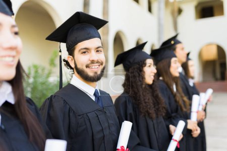 Photo for Latin young man looking at the camera while standing at his graduation ceremony and receiving his college diploma with friends - Royalty Free Image