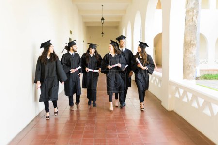 Photo for Cheerful group of graduates walking at the university hall holding their college diploma wearing graduation gowns - Royalty Free Image
