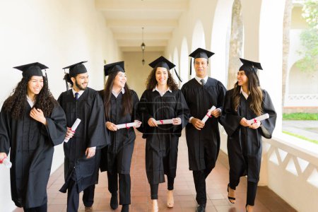 Foto de Emocionado grupo de hombres y mujeres graduados caminando en la sala de la universidad con vestidos de graduación después de recibir su diploma universitario - Imagen libre de derechos