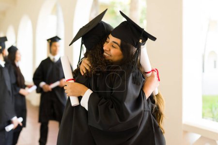 Foto de Mujeres atractivas amigas y graduadas abrazándose y sonriendo viéndose felices durante su graduación universitaria - Imagen libre de derechos
