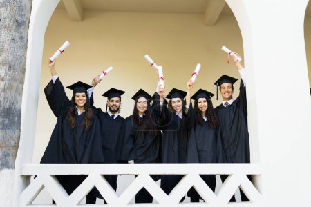 Téléchargez les photos : Portrait d'un groupe d'amis diplômés portant des robes et des casquettes de fin d'études montrant leurs diplômes universitaires au collège - en image libre de droit