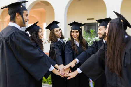 Téléchargez les photos : Groupe souriant de diplômés mettre leurs mains ensemble et célébrer l'université de finition lors de leur cérémonie de remise des diplômes - en image libre de droit