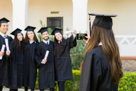 Téléchargez les photos : Jeune femme vue de derrière prenant des photos avec son smartphone de ses amis diplômés pendant l'obtention du diplôme d'université - en image libre de droit