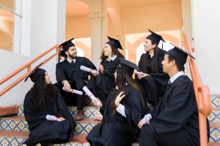 Photo for Cheerful friends sitting on the stairs talking holding their diplomas after their university graduation ceremony - Royalty Free Image