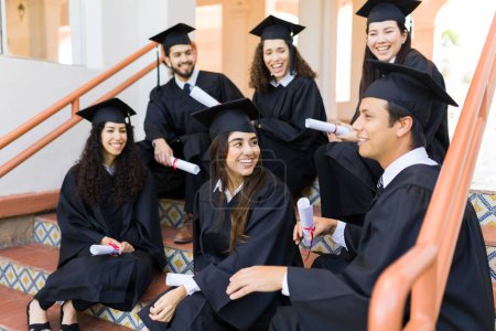 Photo for Attractive graduates friends laughing after their graduation college ceremony while sitting relaxing on the campus stairs - Royalty Free Image