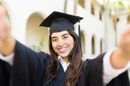 Téléchargez les photos : Belle femme hispanique prenant un selfie avec sa robe de remise des diplômes lors de sa cérémonie sur le campus universitaire - en image libre de droit