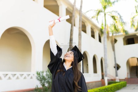 Photo for Beautiful hispanic woman with a graduation gown looking excited while holding her university diploma in the air - Royalty Free Image