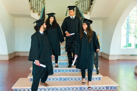 Photo for Happy friends posing standing on the stairs at college campus wearing graduation gowns and diplomas - Royalty Free Image