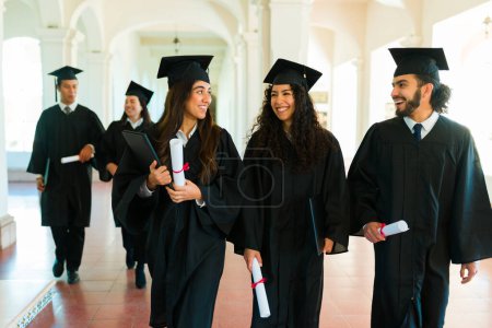 Photo for Beatiful latin women and man friends wearing graduation gowns walking together on university campus after diploma ceremony - Royalty Free Image