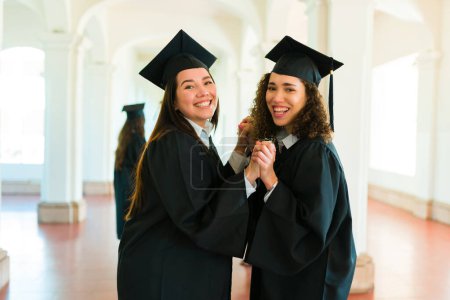 Téléchargez les photos : Femmes meilleures amies avec des robes de remise des diplômes regardant joyeux tenant la main et célébrant obtenir des diplômes universitaires - en image libre de droit