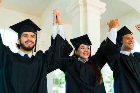 Téléchargez les photos : Amis diplômés à leur cérémonie de remise des diplômes universitaires tenant la main et levant les bras pour célébrer l'obtention de leur diplôme - en image libre de droit