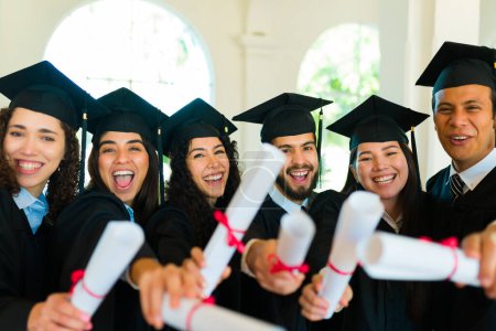 Téléchargez les photos : Groupe excité de diplômés montrant leurs diplômes universitaires et riant lors de leur cérémonie de remise des diplômes - en image libre de droit