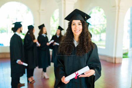 Téléchargez les photos : Magnifique jeune femme avec les cheveux bouclés faisant contact visuel tenant son diplôme universitaire portant une robe de graduation - en image libre de droit