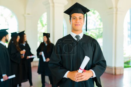 Téléchargez les photos : Beau latin homme qui fait un contact visuel tenant son diplôme universitaire portant une robe de remise des diplômes sur le campus - en image libre de droit
