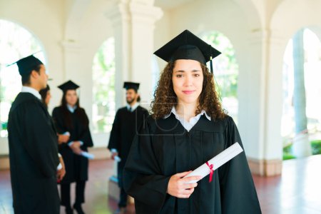 Téléchargez les photos : Fière jeune femme aux cheveux bouclés souriant tenant son diplôme d'études supérieures portant sa robe de graduation et son bonnet - en image libre de droit