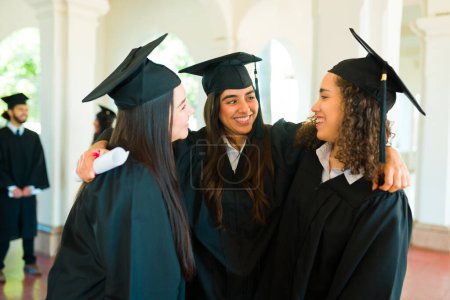 Téléchargez les photos : Femmes gaies meilleurs amis à l'université câlins et parler tout en célébrant le port de robes de graduation - en image libre de droit