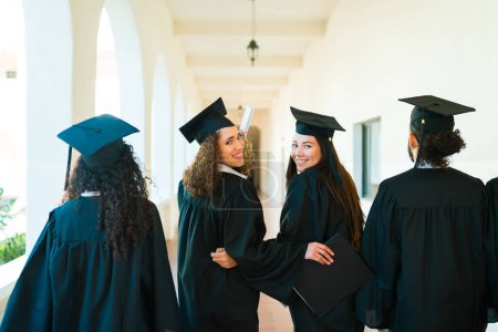 Photo for Beautiful women friends and college graduates seen from behind walking together and smiling making eye contact - Royalty Free Image