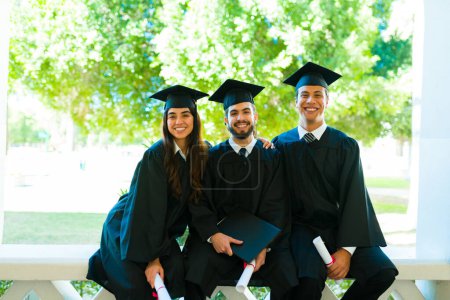 Foto de Attractive friends wearing graduation gowns and caps smiling making eye contact on college campus - Imagen libre de derechos
