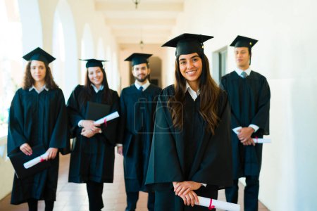 Téléchargez les photos : Bonne femme latine recevant son diplôme d'études collégiales regardant la caméra et souriant avec des amis diplômés sur le campus - en image libre de droit