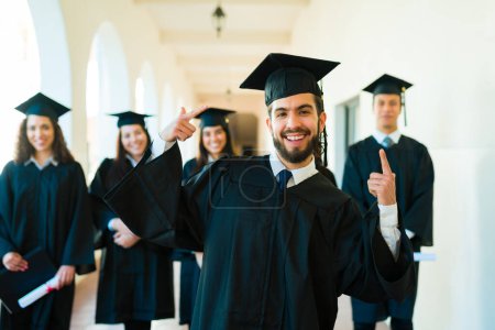 Téléchargez les photos : Heureux attrayant latin homme pointant vers son diplôme robe et casquette tout en souriant sur son campus universitaire - en image libre de droit