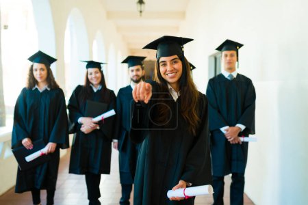 Téléchargez les photos : Magnifique hispanique femme souriant pointant vers l'avant portant une robe de graduation se sentant fière de terminer ses études universitaires - en image libre de droit