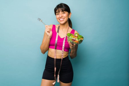 Foto de Atractiva mujer hispana con un estilo de vida de fitness sonriendo mientras disfruta comiendo una deliciosa ensalada saludable para perder peso - Imagen libre de derechos