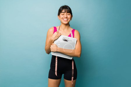 Téléchargez les photos : Femme hispanique regardant gai et heureux tenant une balance atteignant son objectif de perte de poids en faisant un régime alimentaire et séance d'entraînement - en image libre de droit