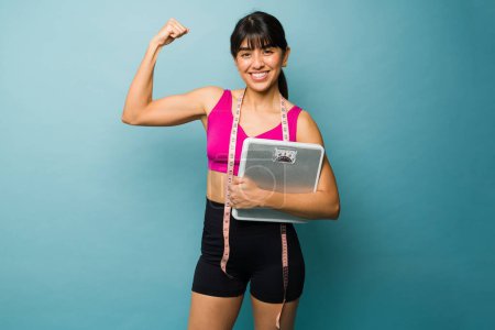 Téléchargez les photos : Forte femme en forme faisant une boucle biceps montrant son corps athlétique après l'exercice et la perte de poids à l'aide d'une balance - en image libre de droit