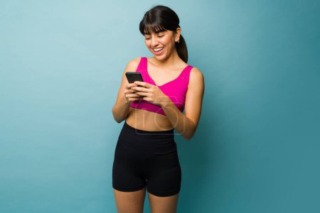 Foto de Hermosa mujer joven en forma riendo mientras envía mensajes en el teléfono inteligente antes de sus ejercicios de entrenamiento - Imagen libre de derechos