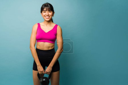 Foto de Mujer atractiva hispana sonriendo haciendo contacto visual mientras usa un kettlebell y levantando pesas durante su entrenamiento - Imagen libre de derechos