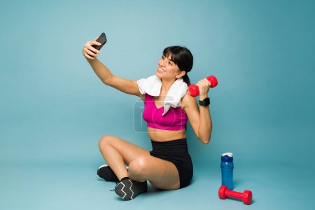 Téléchargez les photos : Coupe sportive jeune femme souriant prendre un selfie avec un smartphone tout en s'entraînant avec haltères poids - en image libre de droit