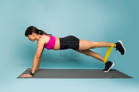 Téléchargez les photos : Vue latérale d'une jeune femme sportive faisant des exercices de planche à l'aide d'une bande de résistance et d'un tapis d'exercice pendant son entraînement - en image libre de droit