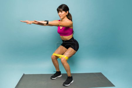 Téléchargez les photos : Superbe jeune femme mexicaine faisant de l'exercice squats et en utilisant une bande de résistance sur un fond bleu - en image libre de droit