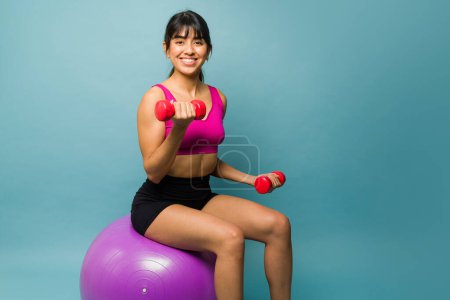 Photo for Gorgeous hispanic fitness woman lifting dumbbell weights and smiling while working out using a stability ball - Royalty Free Image