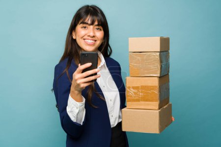 Téléchargez les photos : Magnifique femme latine portant un costume d'affaires professionnel souriant tout en recevant beaucoup de paquets de courrier en utilisant son téléphone - en image libre de droit