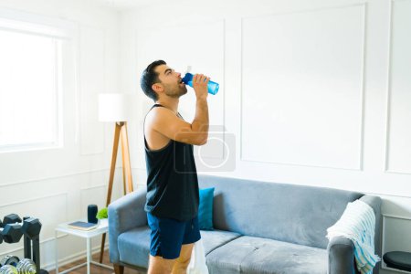 Foto de Sporty fitness man wearing activewear drinking water in the living room before exercising at home - Imagen libre de derechos
