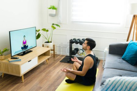Foto de Hombre joven relajándose y haciendo ejercicios de meditación mientras sigue un video de yoga en la sala de estar tv - Imagen libre de derechos