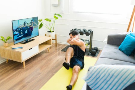 Foto de Deportivo hombre latino haciendo crujidos de bicicleta y ejercicios de cardio mientras ve un video de entrenamiento en casa - Imagen libre de derechos