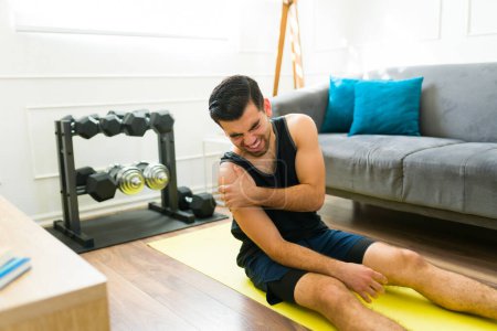 Photo for Pained young man straining his shoulder and suffering an injury after a difficult home workout - Royalty Free Image
