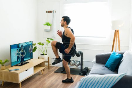 Photo for Sporty young man running in place and doing cardio exercises while watching a workout video on his living room television - Royalty Free Image