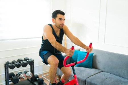 Foto de Hombre latino atractivo haciendo ejercicios de cardio y usando una bicicleta estacionaria durante su entrenamiento - Imagen libre de derechos