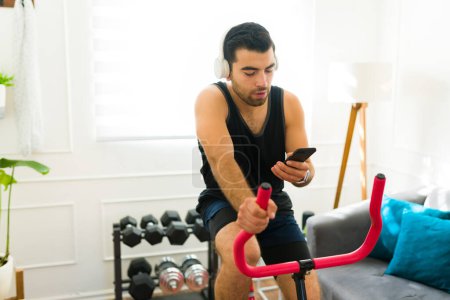 Téléchargez les photos : Handsome sporty man listening to music with headphones and using a fitness app on his phone while exercising with a stationary bicycle - en image libre de droit