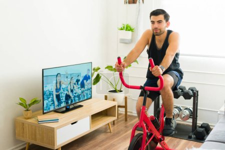 Photo for Determined man with a fitness motivation riding his stationary bicycle and doing cardio exercise at home - Royalty Free Image