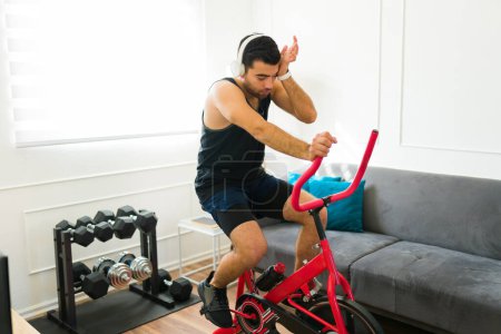 Foto de Hombre latino cansado sudando y sintiendo calor mientras monta la bicicleta estacionaria en la sala de estar y haciendo ejercicio - Imagen libre de derechos