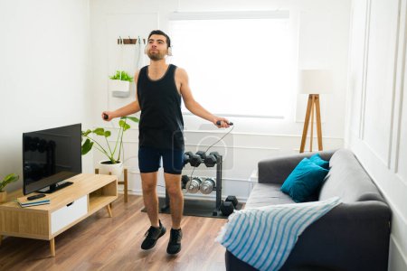 Foto de Active hispanic man with headphones jumping rope and doing his cardio workout in the living room - Imagen libre de derechos
